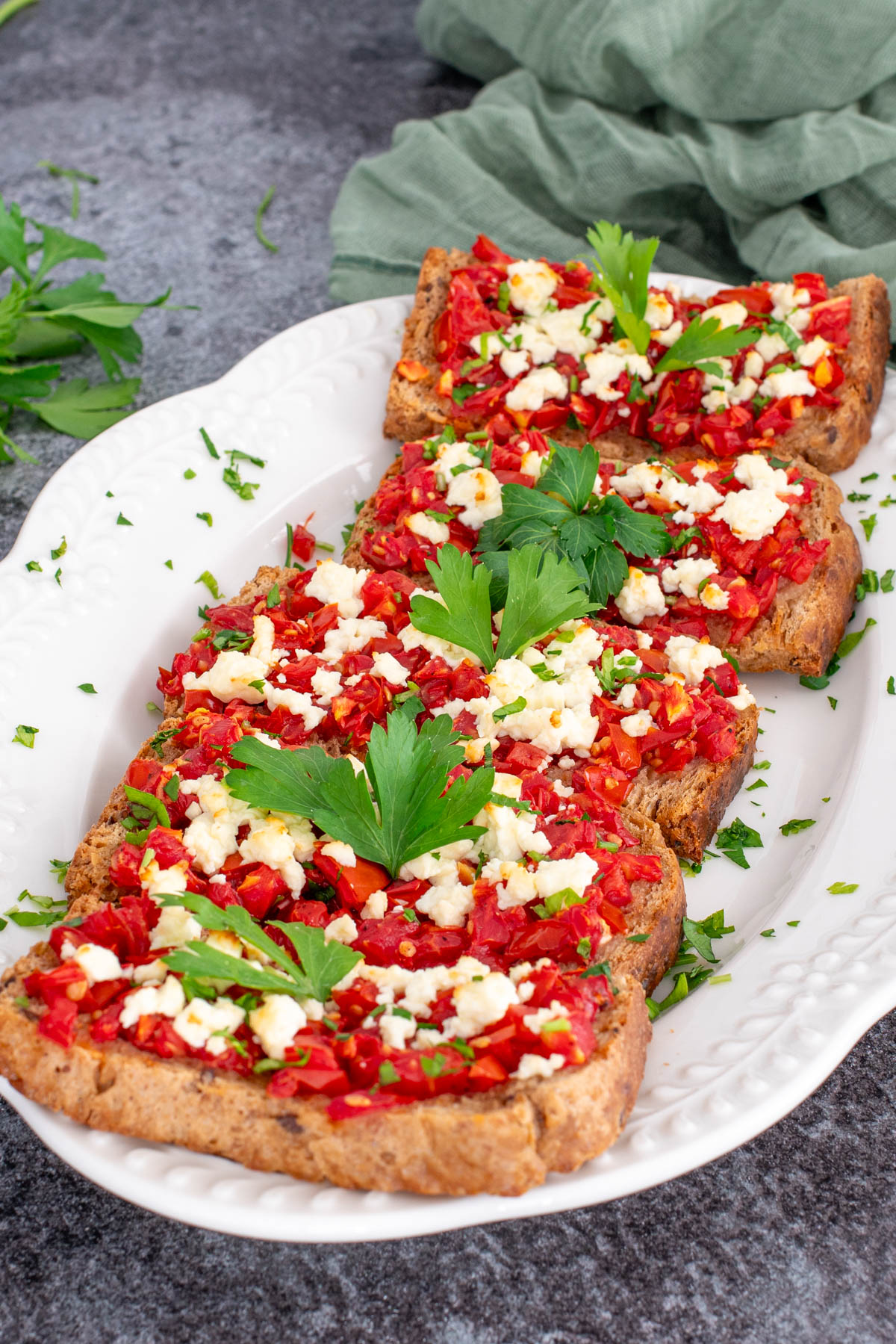 Bruschete coapte cu rosii, branza feta si ulei de masline, asezate pe un platou alb si asezonate cu frunze de patrunjel.