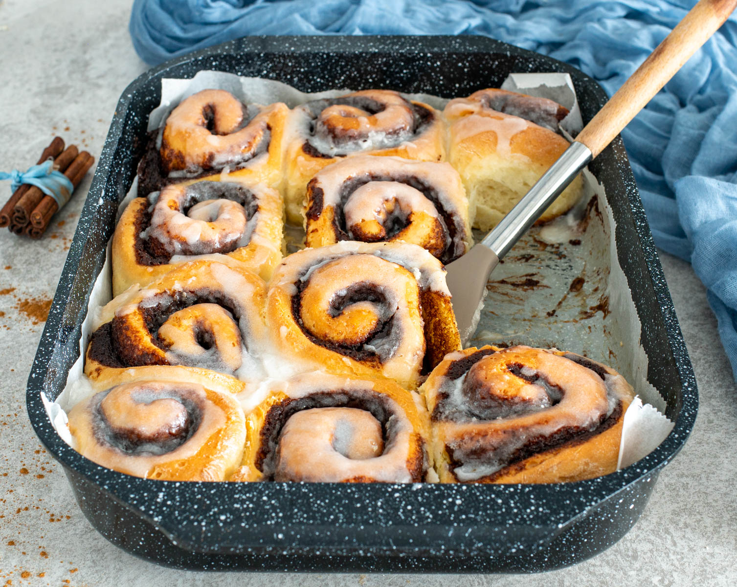 Tava plină cu rulouri pufoase de scorțișoară glazurate, proaspăt scoase din cuptor, cu o spatulă care ridică un rulou și batoane de scorțișoară în fundal.