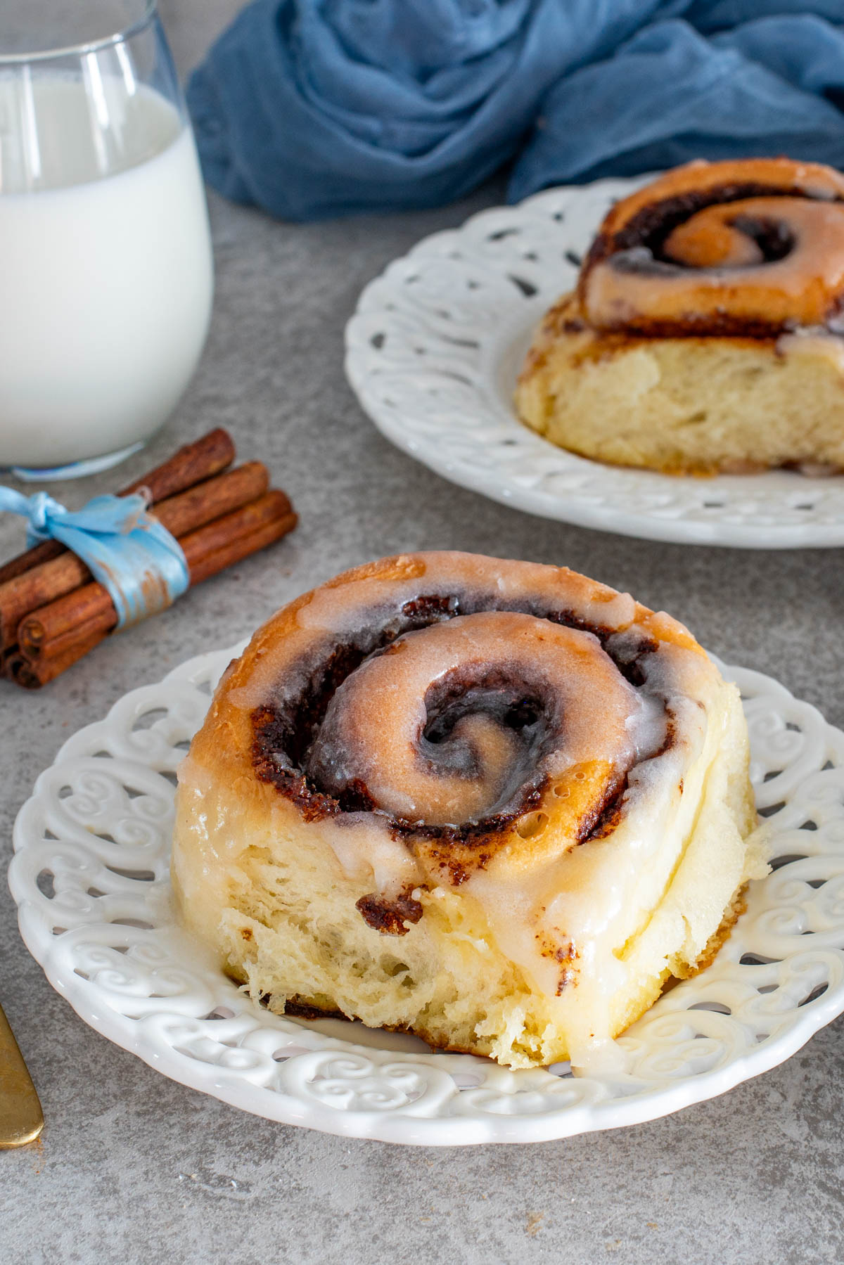 Rulou de scorțișoară glazurat, întreg, așezat pe o farfurie albă decorată, cu un pahar de lapte și batoane de scorțișoară în fundal.