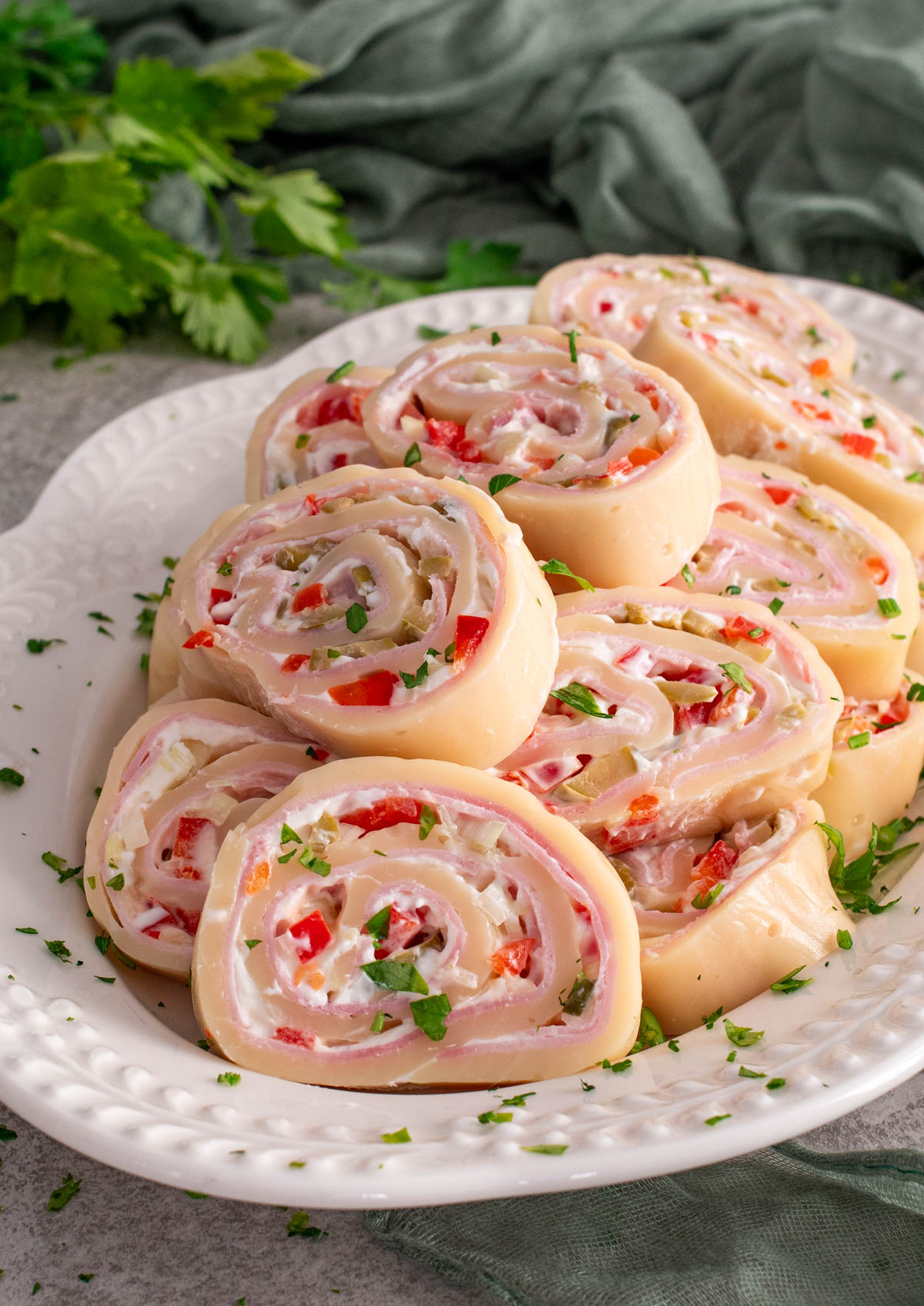 Felii de ruladă de cașcaval umplută cu șuncă, cremă de br&acirc;nză și legume tocate (ardei și castraveți), aranjate pe o farfurie albă, decorate cu pătrunjel proaspăt, cu un șervet verde și verdeață &icirc;n fundal.