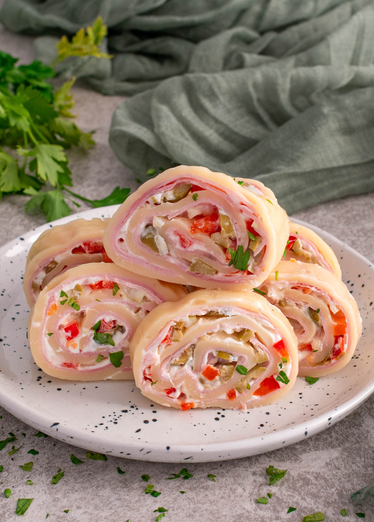 Felii de ruladă de cașcaval umplută cu șuncă, cremă de br&acirc;nză și legume tocate (ardei și castraveți), aranjate pe o farfurie albă, decorate cu pătrunjel proaspăt, cu un șervet verde și verdeață &icirc;n fundal.