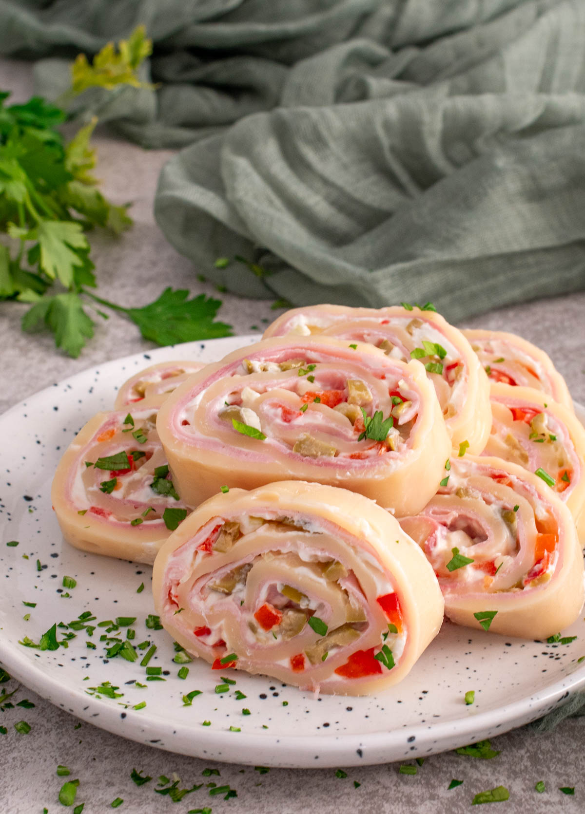 Felii de ruladă de cașcaval umplută cu șuncă, cremă de br&acirc;nză și legume tocate (ardei și castraveți), aranjate pe o farfurie albă, decorate cu pătrunjel proaspăt, cu un șervet verde și verdeață &icirc;n fundal.