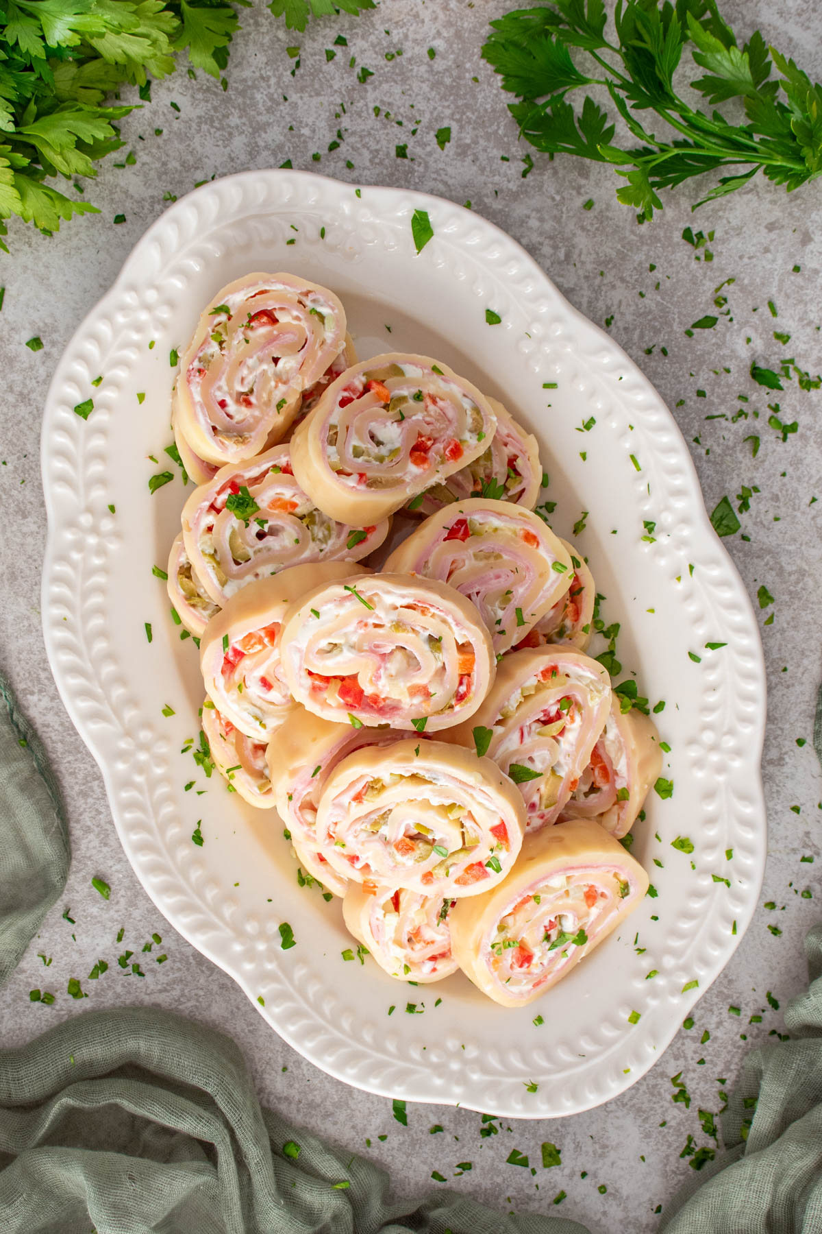 Platou oval alb cu felii de ruladă de cașcaval umplută cu șuncă, cremă de br&acirc;nză și legume tocate, presărate cu pătrunjel verde, fotografiat de sus, cu frunze de pătrunjel și un șervet verde &icirc;n jur.
