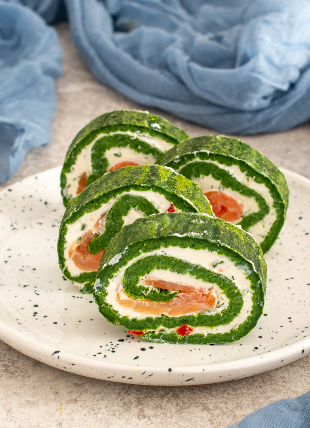 Felii de ruladă cu blat de spanac, cremă de br&acirc;nză și somon afumat, pe farfurie albă.