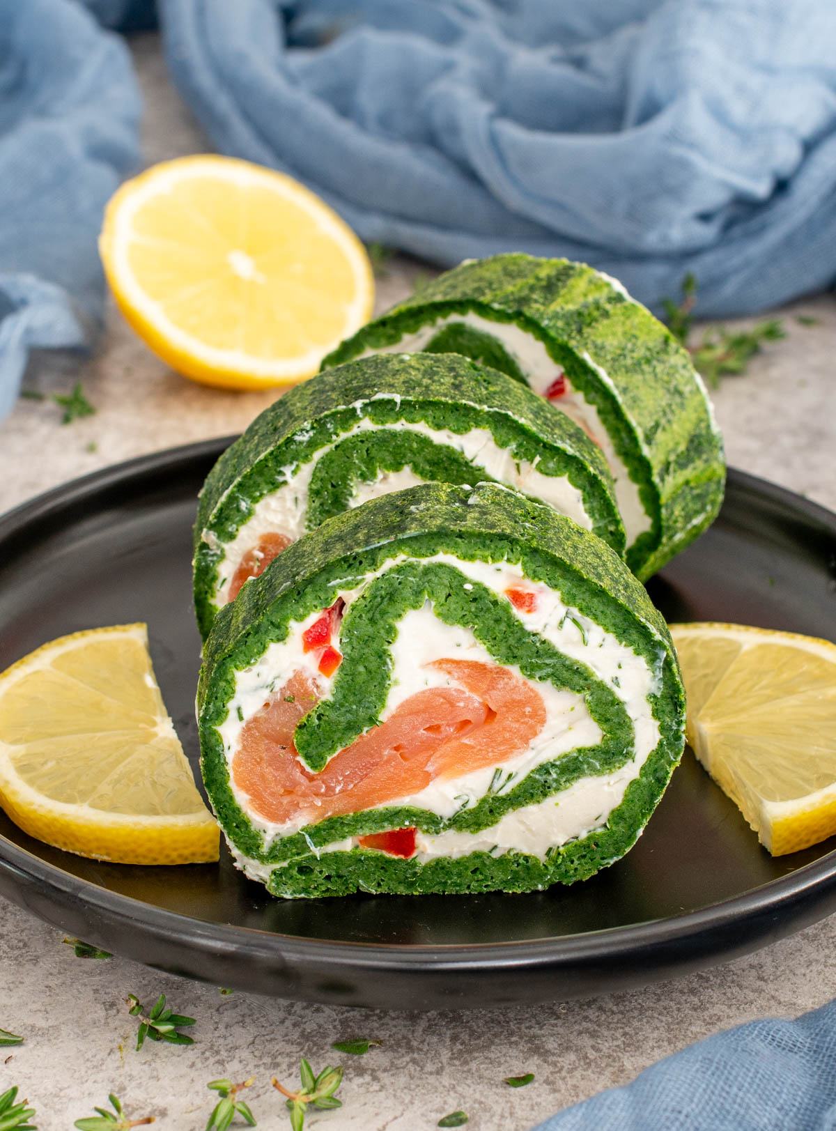 Ruladă de spanac tăiată felii, cu umplutură de cremă de br&acirc;nză, somon afumat și ardei, pe farfurie neagră