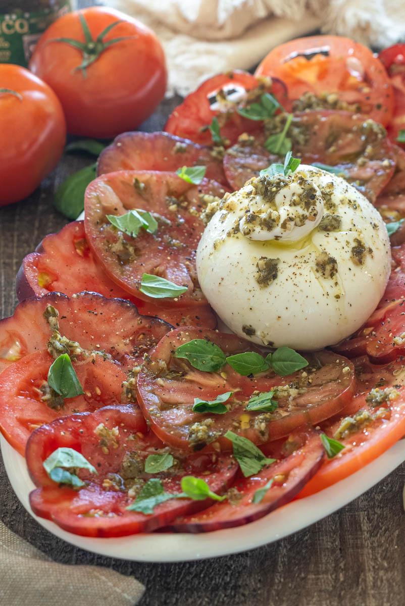 Salată cu burrata cremoasă, roșii feliate, pesto de busuioc, frunze proaspete de busuioc și reducție balsamică, servită pe un platou alb.