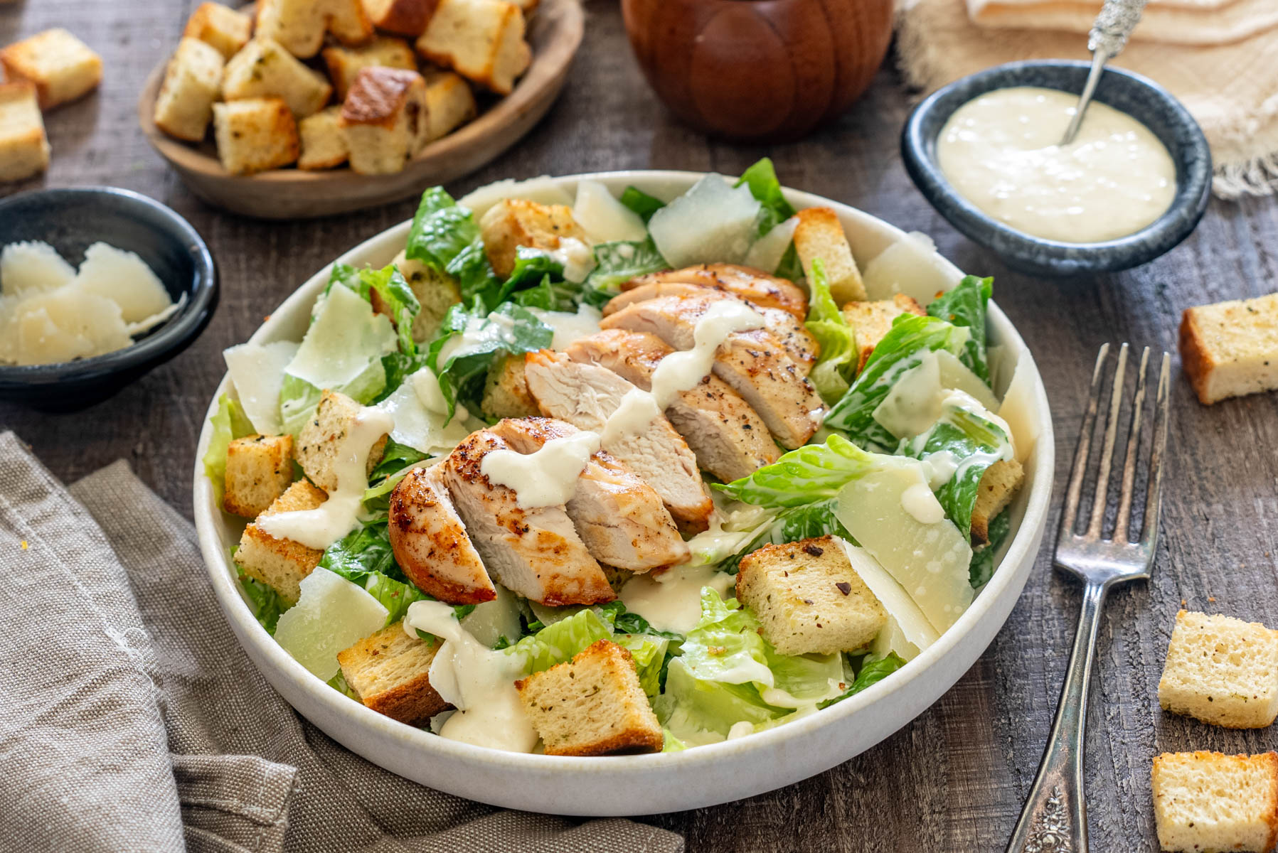 Salată Caesar cu piept de pui la grătar, salată romaine, crutoane crocante, fulgi de parmezan și dressing Caesar, servită într-un bol, alături de dressing și parmezan.