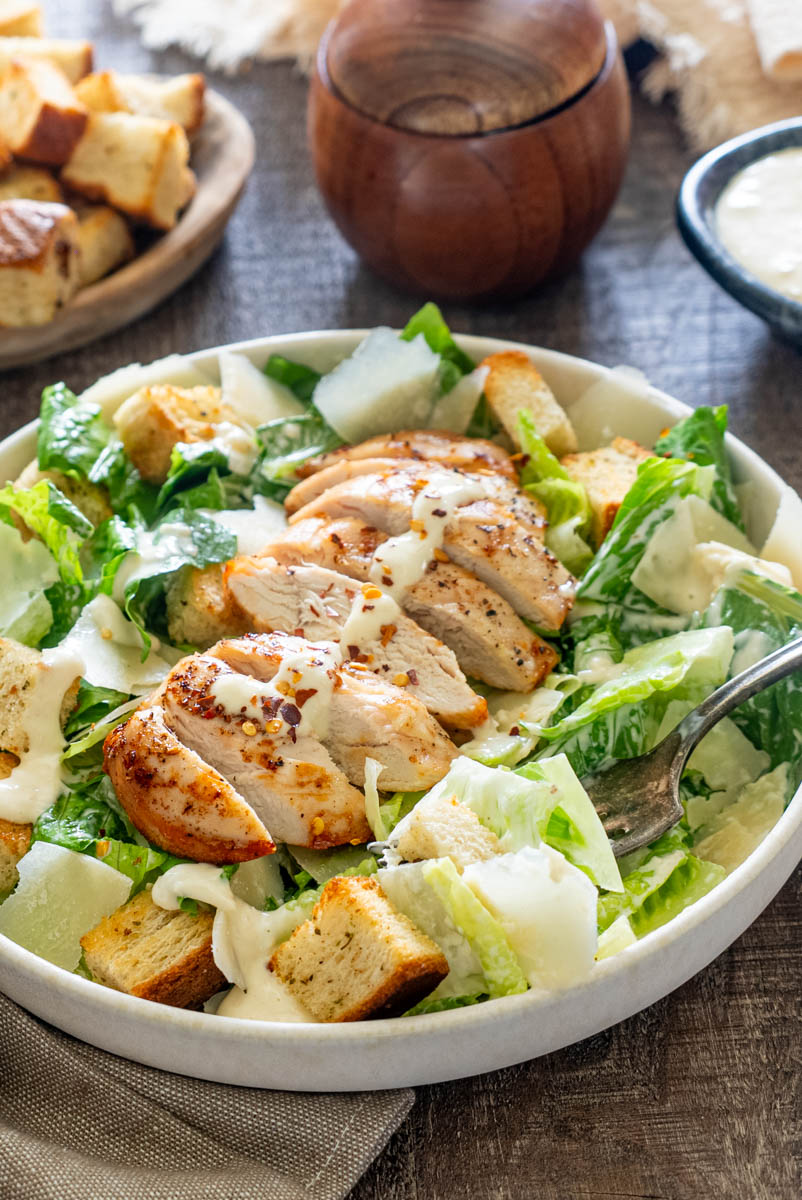 Salată Caesar cu piept de pui la grătar, salată romaine, crutoane, parmezan și dressing cremos, servită într-un bol alb.