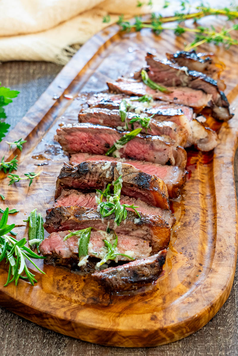 Steak la grătar feliat, gătit medium-rare, servit pe un platou din lemn cu unt aromatizat, usturoi și ierburi proaspete.
