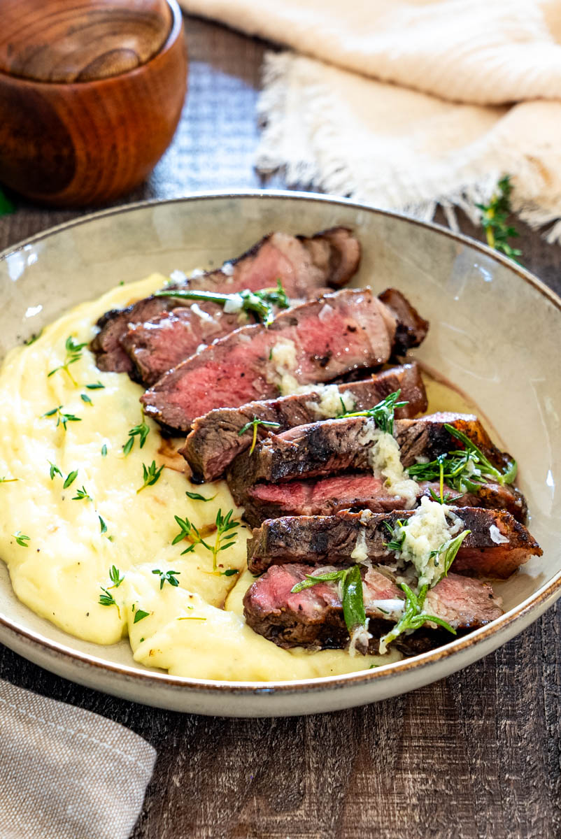 Steak la grătar feliat, servit peste piure cremos de cartofi cu unt aromatizat, usturoi și ierburi proaspete.