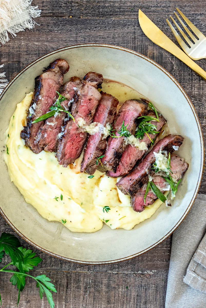Steak de vită fraged cu piure de cartofi și unt aromatizat, servit într-un bol rustic.