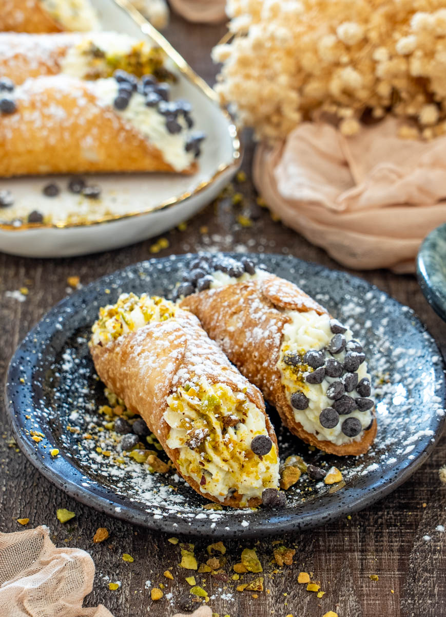 Cannoli crocanți umpluți cu cremă de ricotta, decorați cu fistic și fulgi de ciocolată, serviți pe un platou.