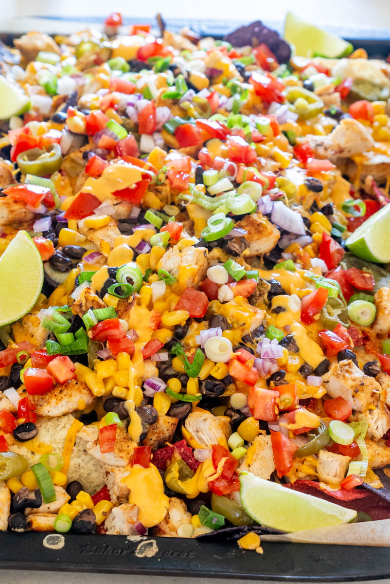 Tavă cu nachos cu pui, acoperiți cu br&acirc;nză cheddar topită, fasole neagră, porumb, roșii, ceapă roșie, ceapă verde, jalape&ntilde;o și felii de lime.