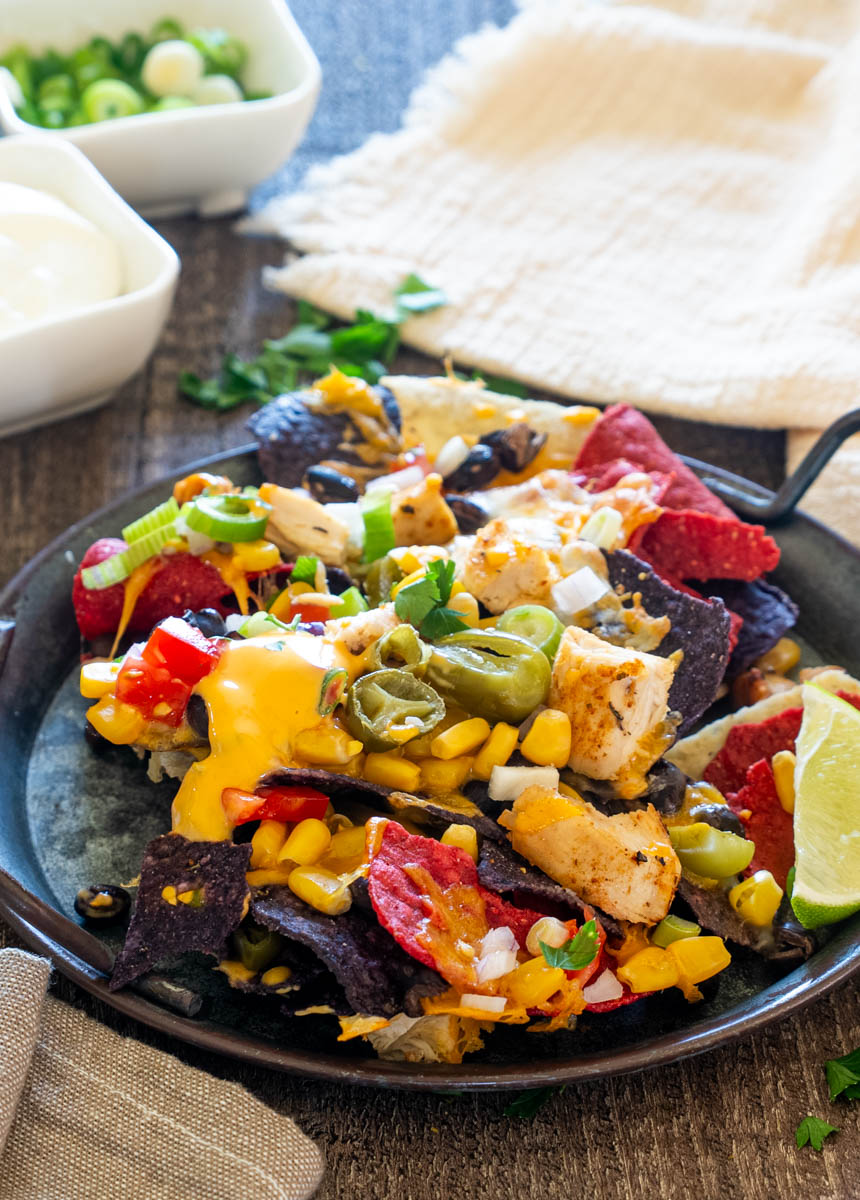 Nachos cu pui, br&acirc;nză cheddar topită, jalape&ntilde;o, porumb, fasole neagră și ceapă verde, serviți cu felie de lime.