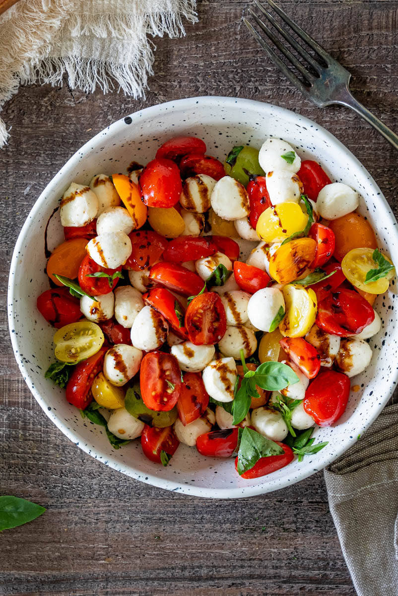 Salată Caprese cu roșii cherry roșii și galbene, mini mozzarella, frunze de busuioc proaspăt și glazură de oțet balsamic, servită într-un bol.