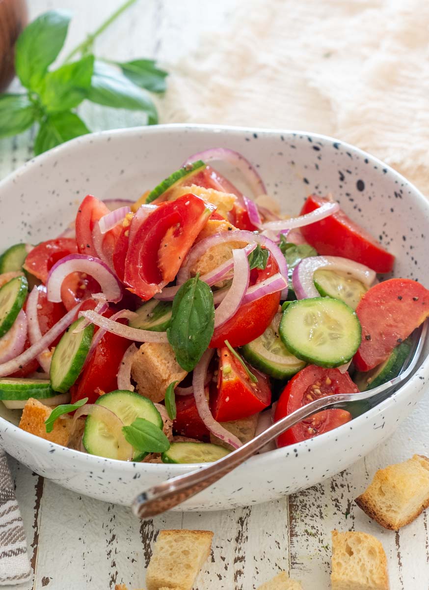 Salată Panzanella cu roșii, castraveți, ceapă roșie, bucăți de pâine și busuioc, într-un bol alb.