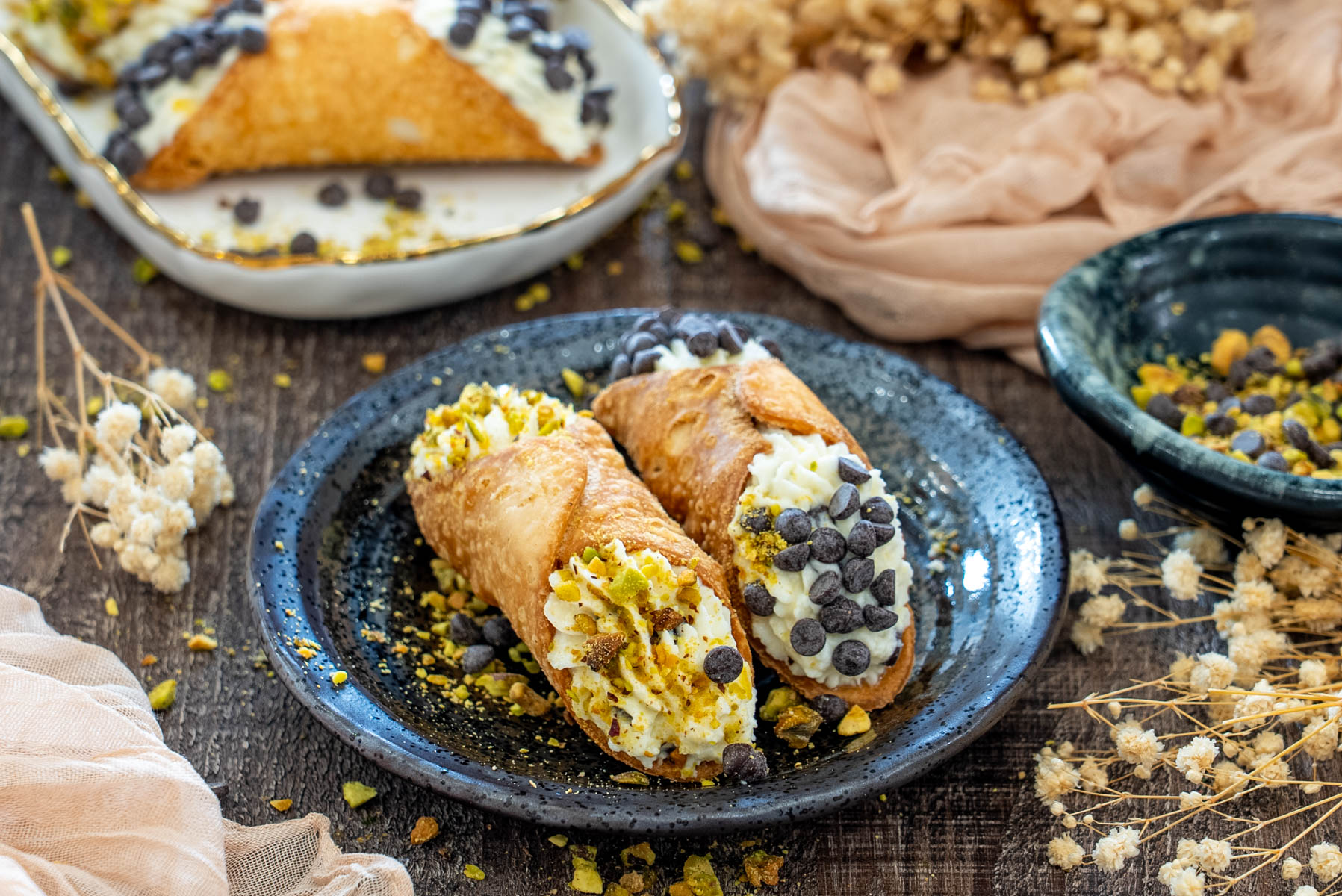 Două cannoli italienești cu umplutură cremoasă, unul decorat cu fistic și celălalt cu chipsuri de ciocolată, servite pe farfurie.
