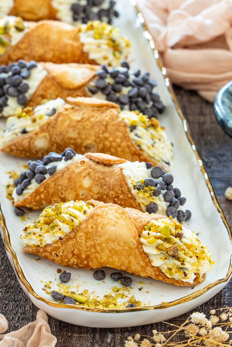 Cannoli crocanți umpluți cu cremă fină de ricotta, cu fistic mărunțit și fulgi de ciocolată, aranjați pe un platou.