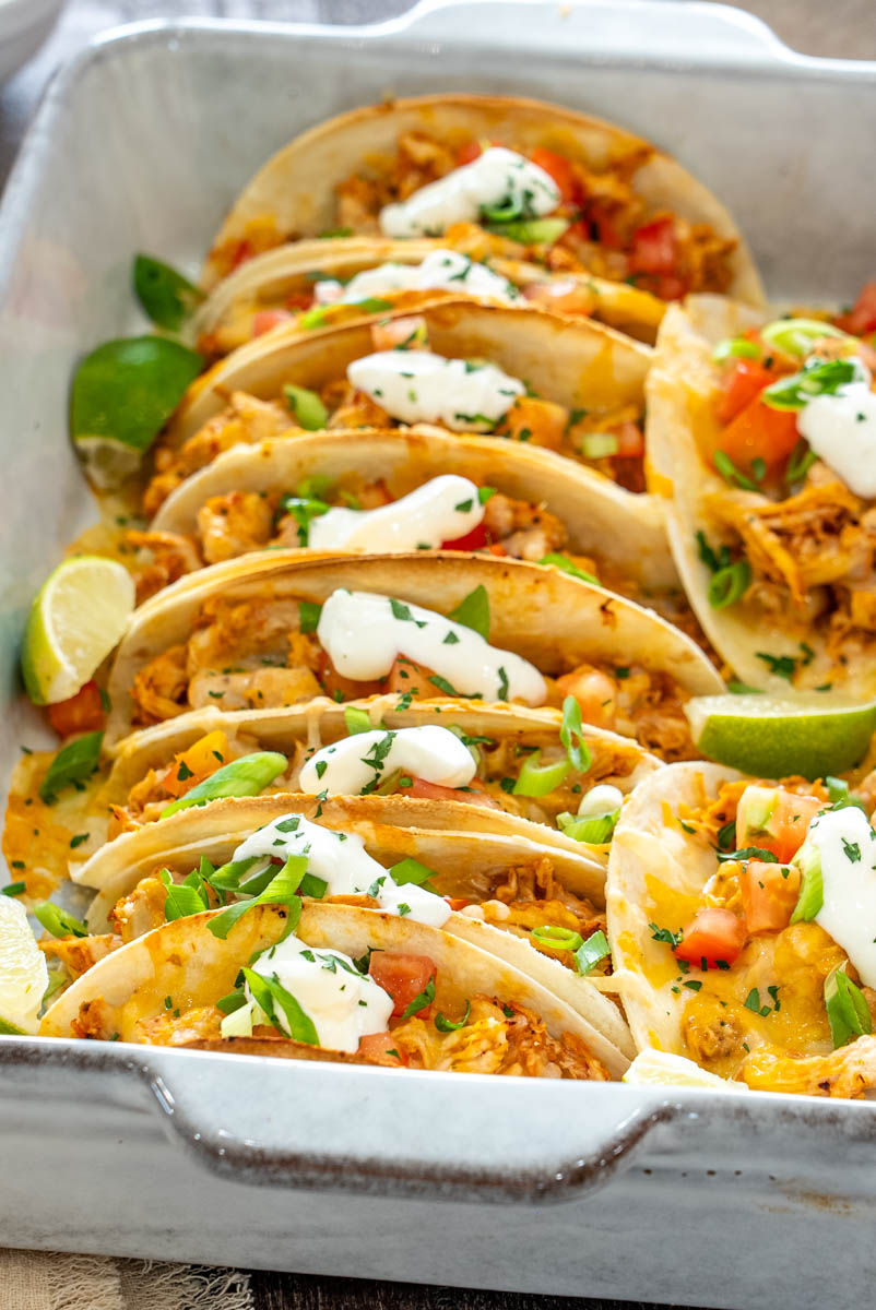 Tacos cu pui, br&acirc;nză topită, roșii, ceapă verde și sos cremos, serviți cu felii de lime.