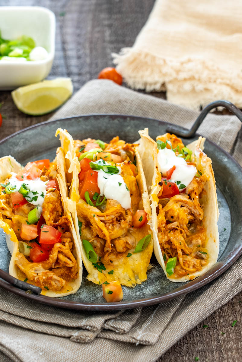 Tacos cu pui și br&acirc;nză la cuptor, garnisiți cu roșii, ceapă verde, sos cremos și lime proaspăt.
