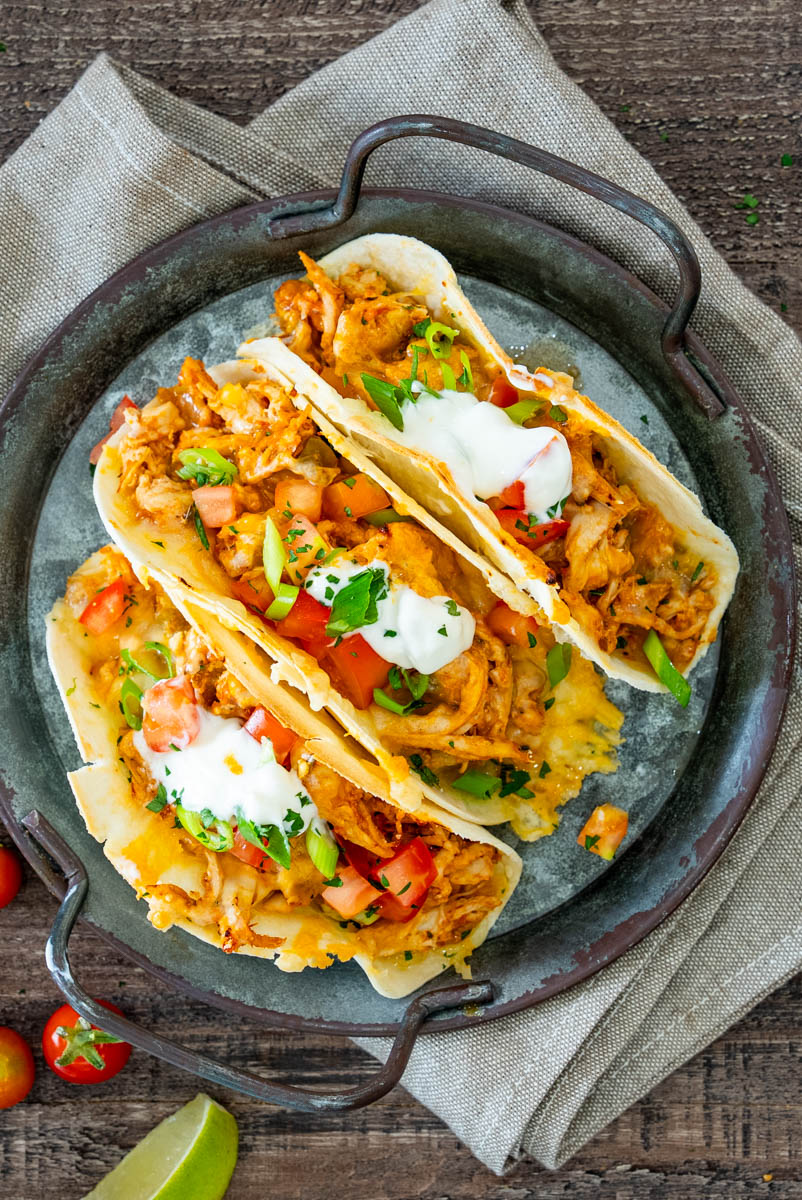 Tacos cu pui și br&acirc;nză topită, umpluți cu roșii proaspete și ceapă verde, serviți cu sos cremos și lime.