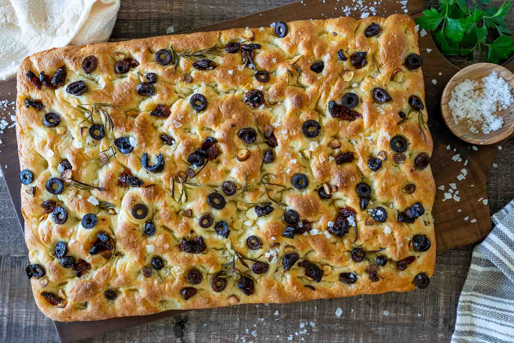 Focaccia întreagă cu măsline negre, roșii uscate și rozmarin, văzută de sus