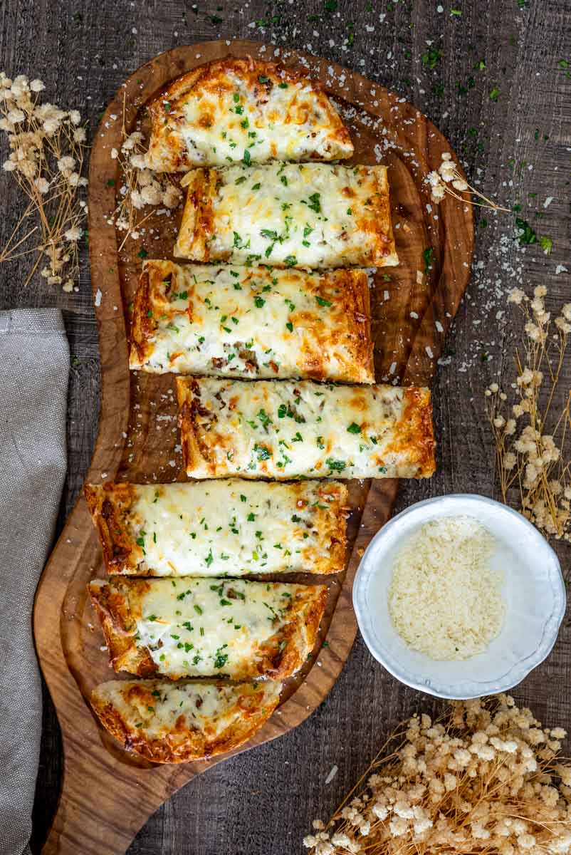 Felii de p&acirc;ine cu usturoi, mozzarella topită și parmezan, servite pe platou de lemn.