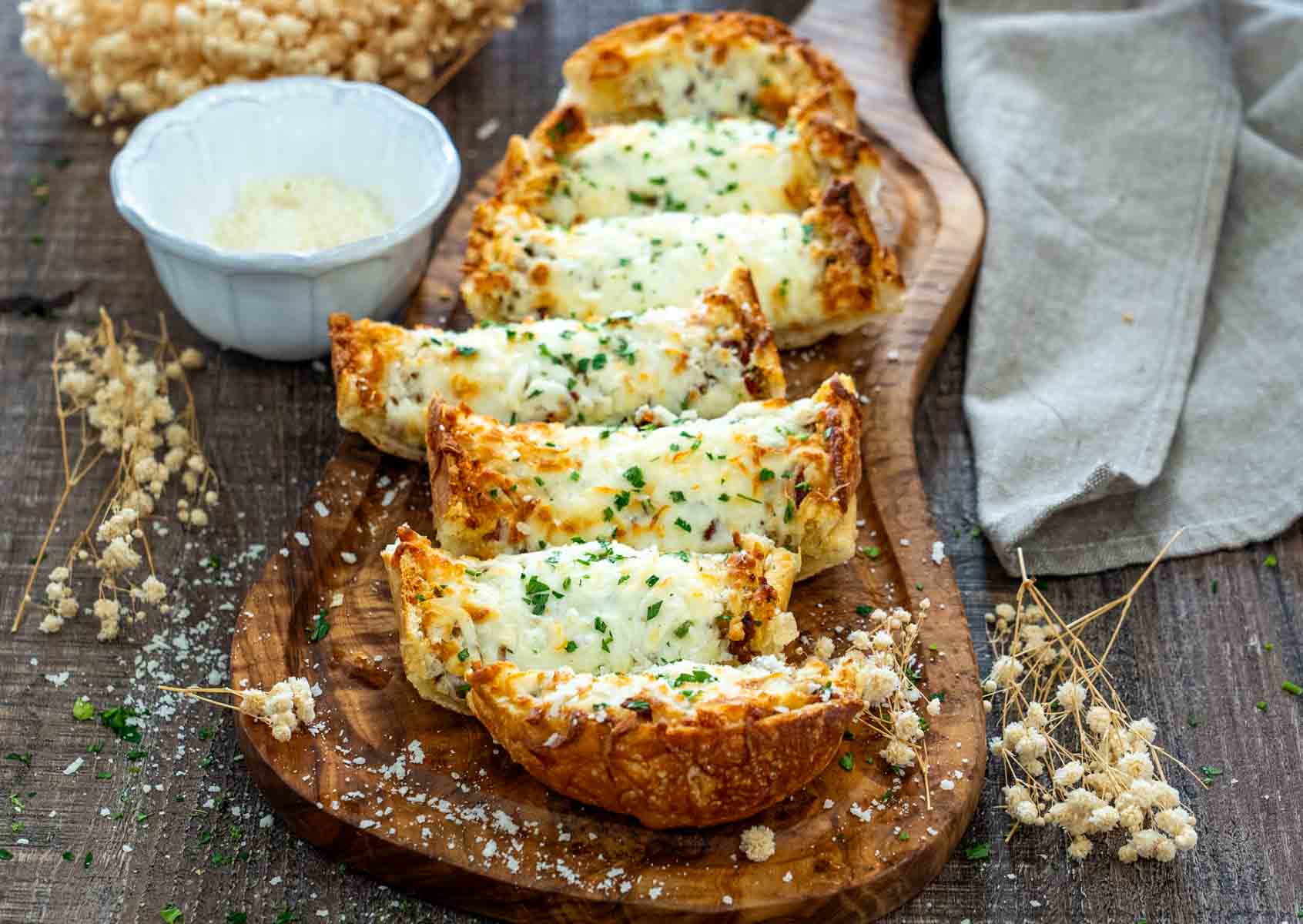 Felii de p&acirc;ine cu usturoi, mozzarella topită și pătrunjel, servite pe platou de lemn.