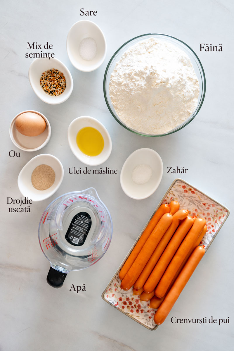 Ingrediente pentru covridogi de casă: făină, drojdie uscată, apă, ulei de măsline, zahăr, sare, ou, crenvurști de pui și mix de semințe, cu denumirile indicate &icirc;n fotografie.