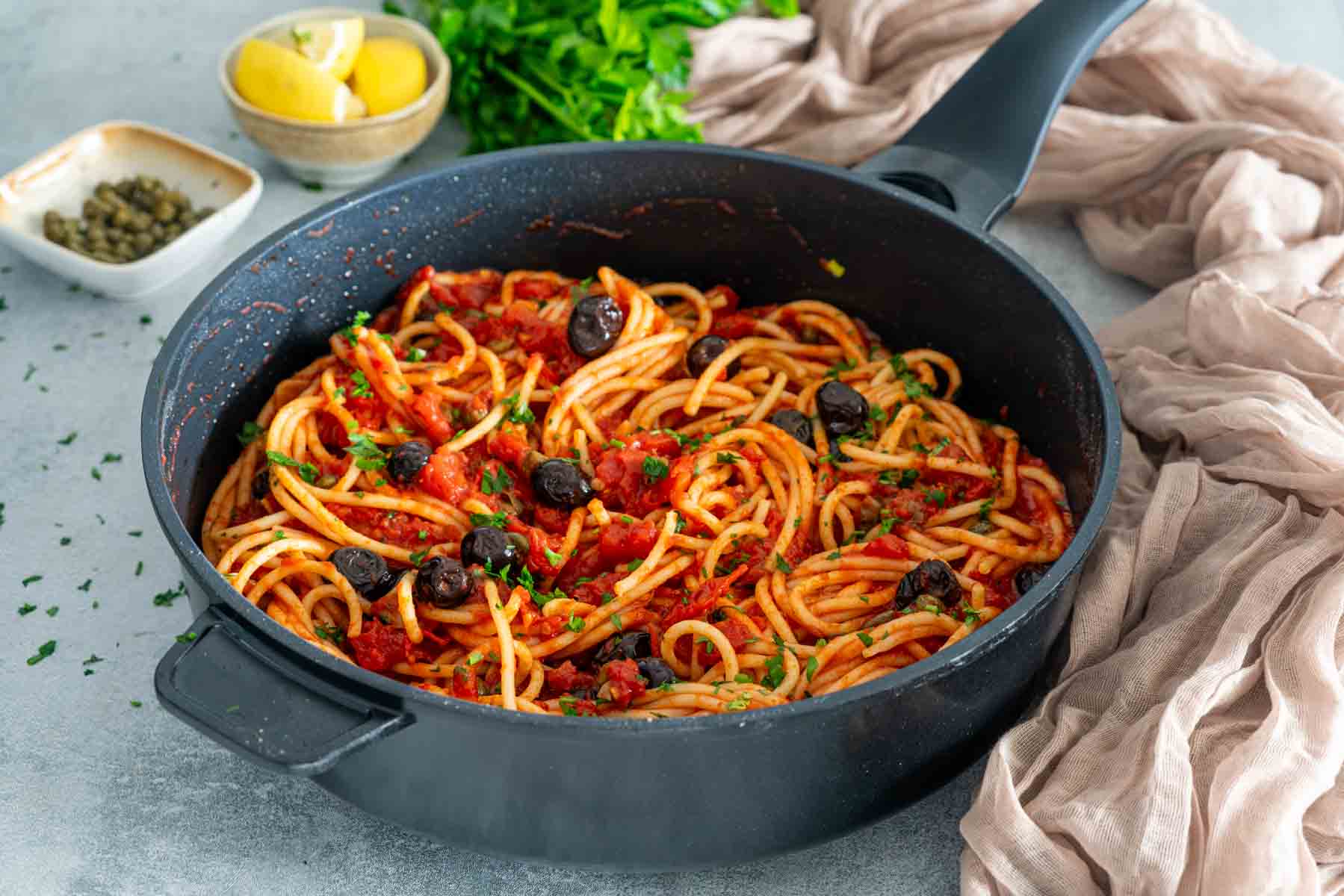 Paste spaghetti Putanesca cu sos de roșii, măsline negre și pătrunjel, servite &icirc;ntr-o tigaie neagră.