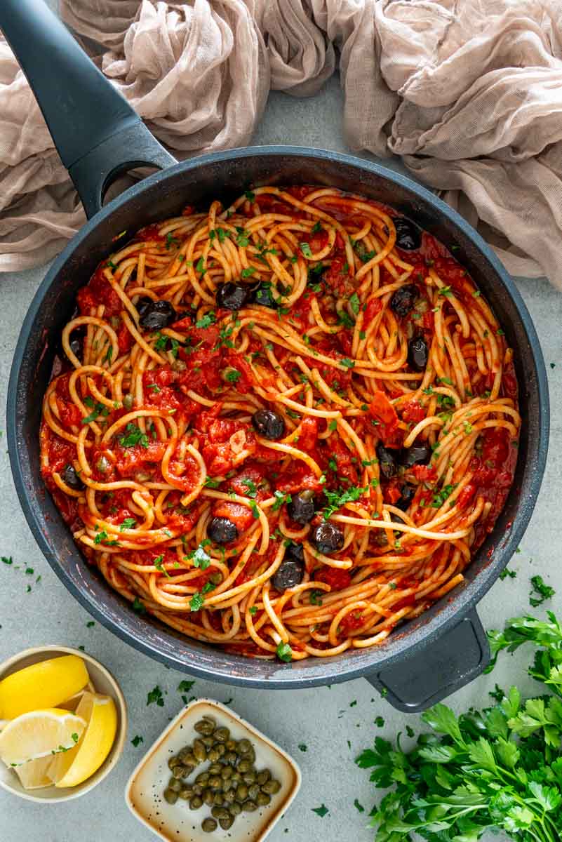 Paste spaghetti Putanesca cu sos de roșii, măsline negre, capere și pătrunjel proaspăt, pregătite &icirc;ntr-o tigaie.
