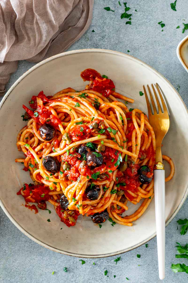 Porție de paste Putanesca cu spaghetti, sos de roșii, măsline negre, capere și pătrunjel proaspăt, servită &icirc;ntr-o farfurie ceramică.