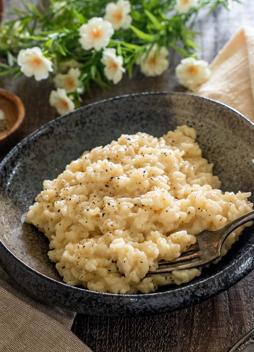 Risotto cu parmezan și piper negru într-un bol gri, cu o furculiță în prim-plan și flori albe în fundal.