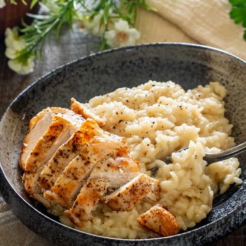 Risotto cu parmezan și felii de piept de pui rumenit, într-un bol închis la culoare, cu o furculiță care ridică orezul cremos.
