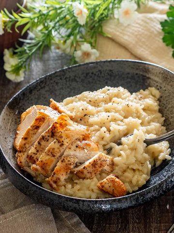 Risotto cu parmezan și felii de piept de pui rumenit, într-un bol închis la culoare, cu o furculiță care ridică orezul cremos.