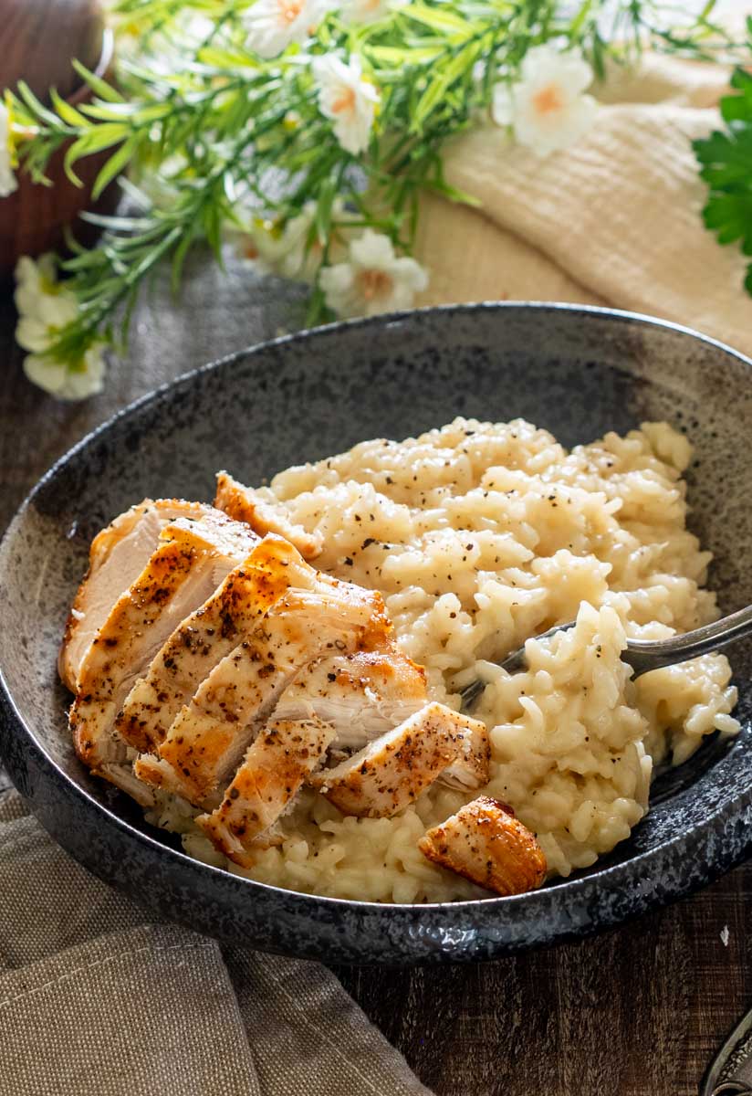 Risotto cu parmezan și felii de piept de pui rumenit, într-un bol închis la culoare, cu o furculiță care ridică orezul cremos.