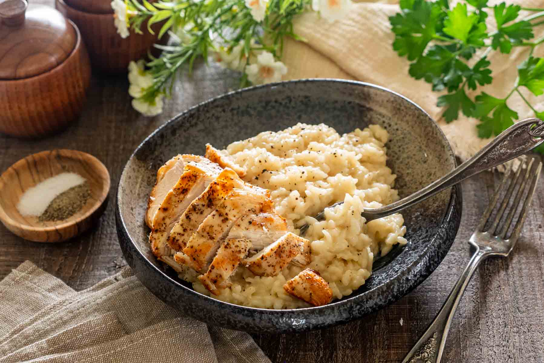 Bol gri cu risotto cu parmezan, felii de piept de pui rumenit și o furculiță cu orez, pe o masă rustică.