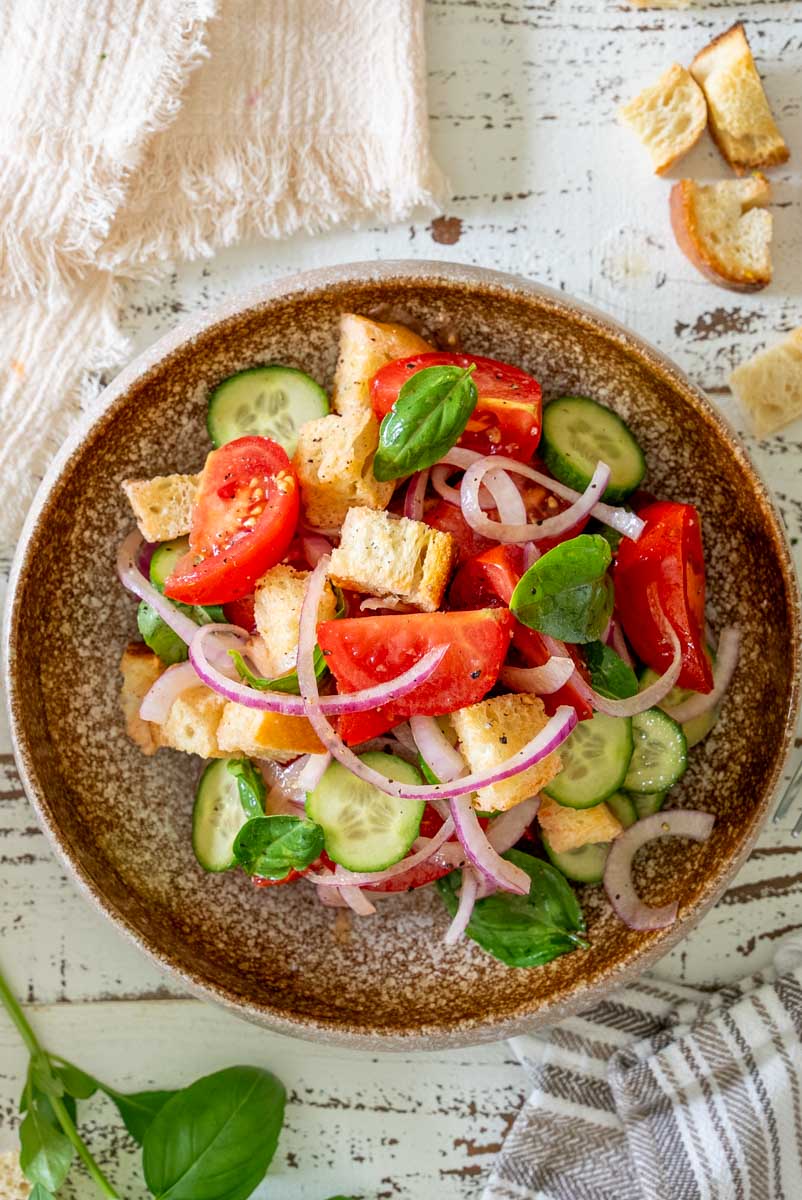Salată Panzanella într-un bol alb, cu roșii, felii de castravete, ceapă roșie, pâine rumenită și frunze de busuioc.