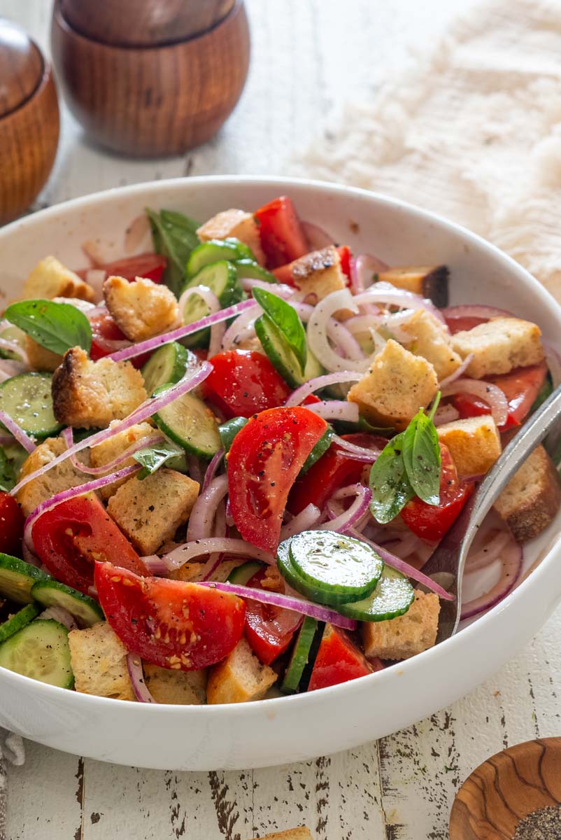 Salată Panzanella într-un bol alb, cu roșii, felii de castravete, ceapă roșie, pâine rumenită și frunze de busuioc.