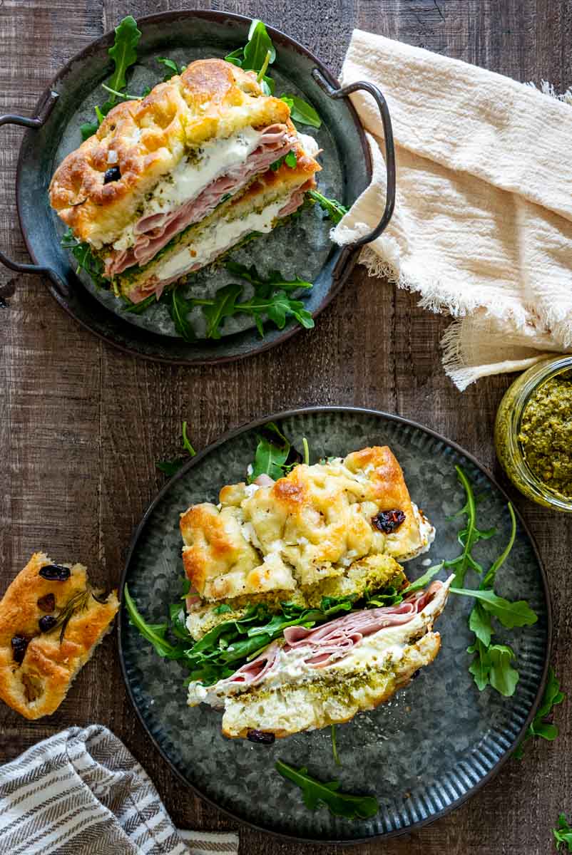Sandvișuri italiene cu focaccia, mortadella, br&acirc;nză cremoasă, pesto și rucola, pe două farfurii &icirc;nchise la culoare, fotografiate de sus.