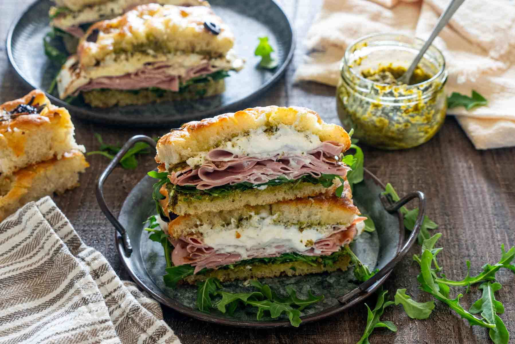 Două jumătăți de sandviș italian cu focaccia, mortadella, br&acirc;nză cremoasă, pesto și rucola, așezate una peste alta pe o tavă metalică.