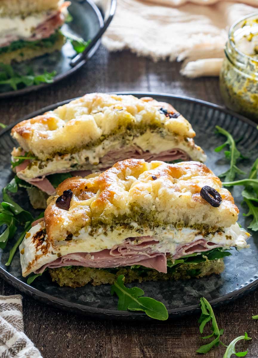 Două porții de sandviș italian cu focaccia cu măsline, mortadella, br&acirc;nză cremoasă, pesto și rucola, pe o farfurie neagră.