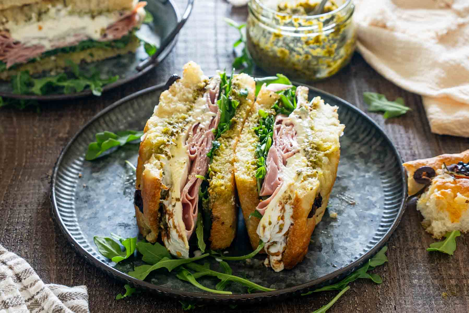 Sandviș italian cu focaccia, mortadella, br&acirc;nză cremoasă, pesto și rucola, tăiat &icirc;n două