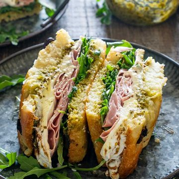 Sandviș cu focaccia, mortadella, br&acirc;nză cremoasă, pesto și rucola, tăiat &icirc;n jumătăți pe o farfurie neagră.