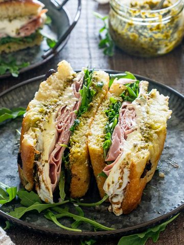 Sandviș cu focaccia, mortadella, br&acirc;nză cremoasă, pesto și rucola, tăiat &icirc;n jumătăți pe o farfurie neagră.