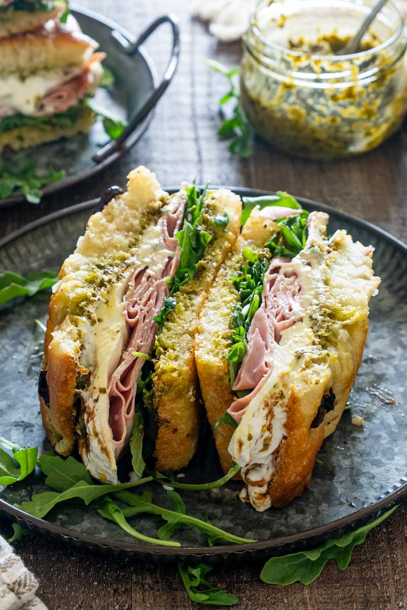 Sandviș cu focaccia, mortadella, br&acirc;nză cremoasă, pesto și rucola, tăiat &icirc;n jumătăți pe o farfurie neagră.